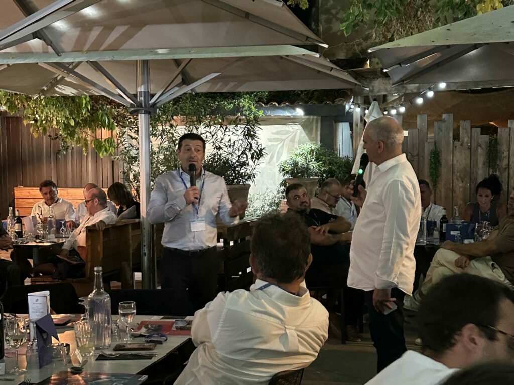 Deux hommes debout échangent au micro devant les participants attablés sur la terrasse de La Pergola, lors de la rentrée économique de La Mêlée Ouverte.